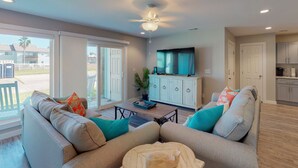 A spacious living room with a large sofa, a coffee table, and a television on a cabinet. There is a sliding glass door leading to a patio area. - 1st Floor, Shared Pool and Hot Tub, Near Marina and Ferry (Port Aransas)