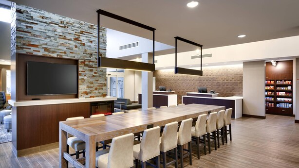 A modern kitchen with a long wooden table, bar stools, a built-in TV, and a brick accent wall. - Residence Inn by Marriott San Marcos (San Marcos)
