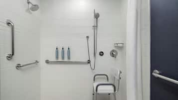 A shower area with white tiled walls, a handheld showerhead, a handheld shower attachment, a grab bar, a seat, and two bottles of lotion.