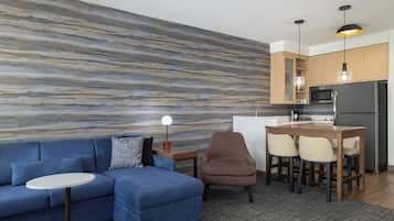 Een moderne woonkamer met een blauwe bank, een kleine ronde tafel, een bar met krukken en een keuken met houten kastjes en roestvrijstalen apparatuur.