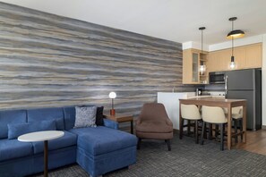 A modern living room with a blue sofa, a small round table, a bar area with stools, and a kitchen with wooden cabinets and stainless steel appliances. - Residence Inn by Marriott San Marcos (San Marcos)