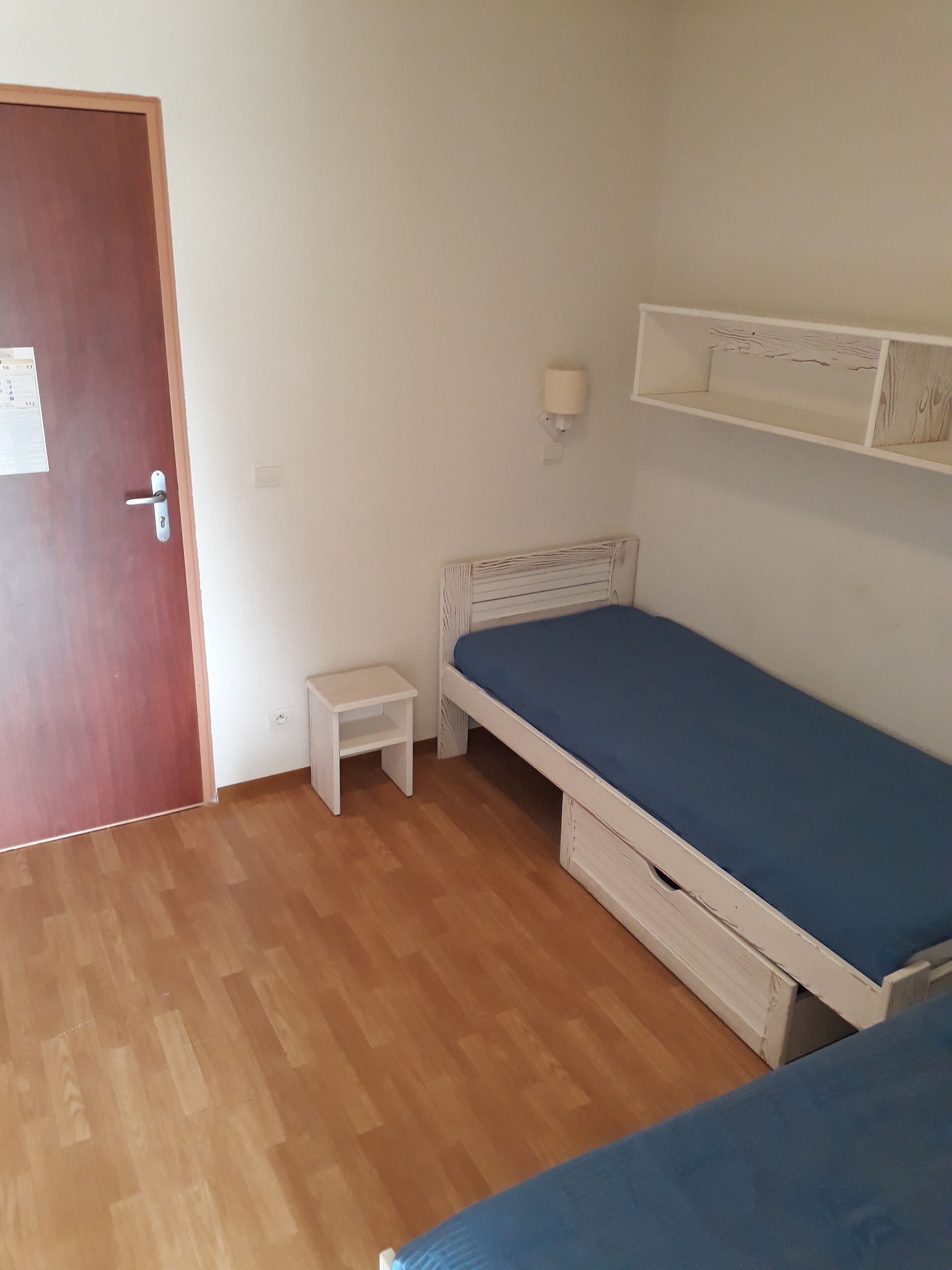 Une pièce avec un plancher en bois, un lit avec un matelas bleu, une petite table de chevet blanche, une étagère blanche et une porte en bois.