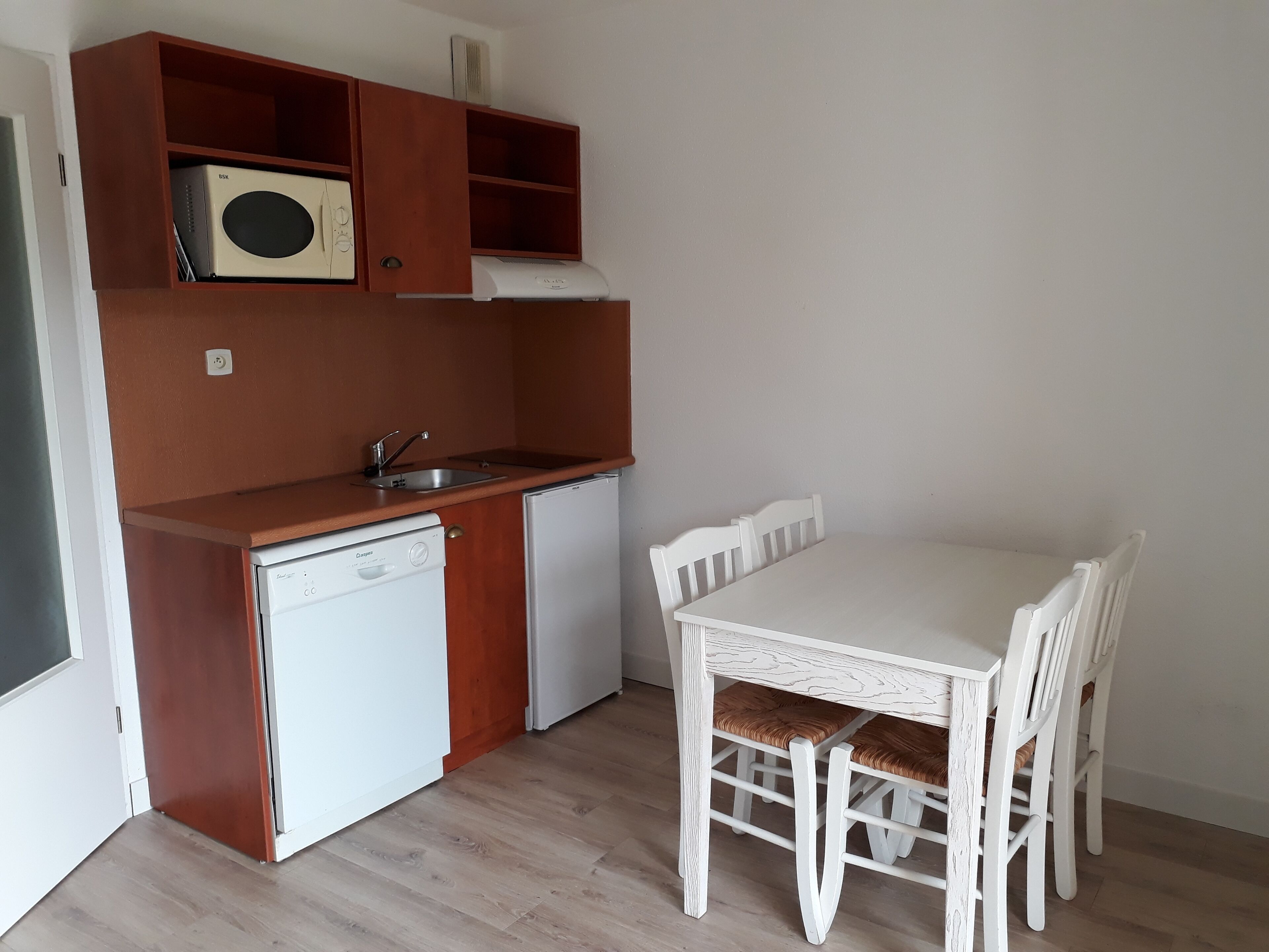 A compact kitchen with a dishwasher, a sink, and a microwave on a wooden shelf. There is a white table with four chairs and a white cabinet below.