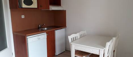 A compact kitchen with a dishwasher, a sink, and a microwave on a wooden shelf. There is a white table with four chairs and a white cabinet below.