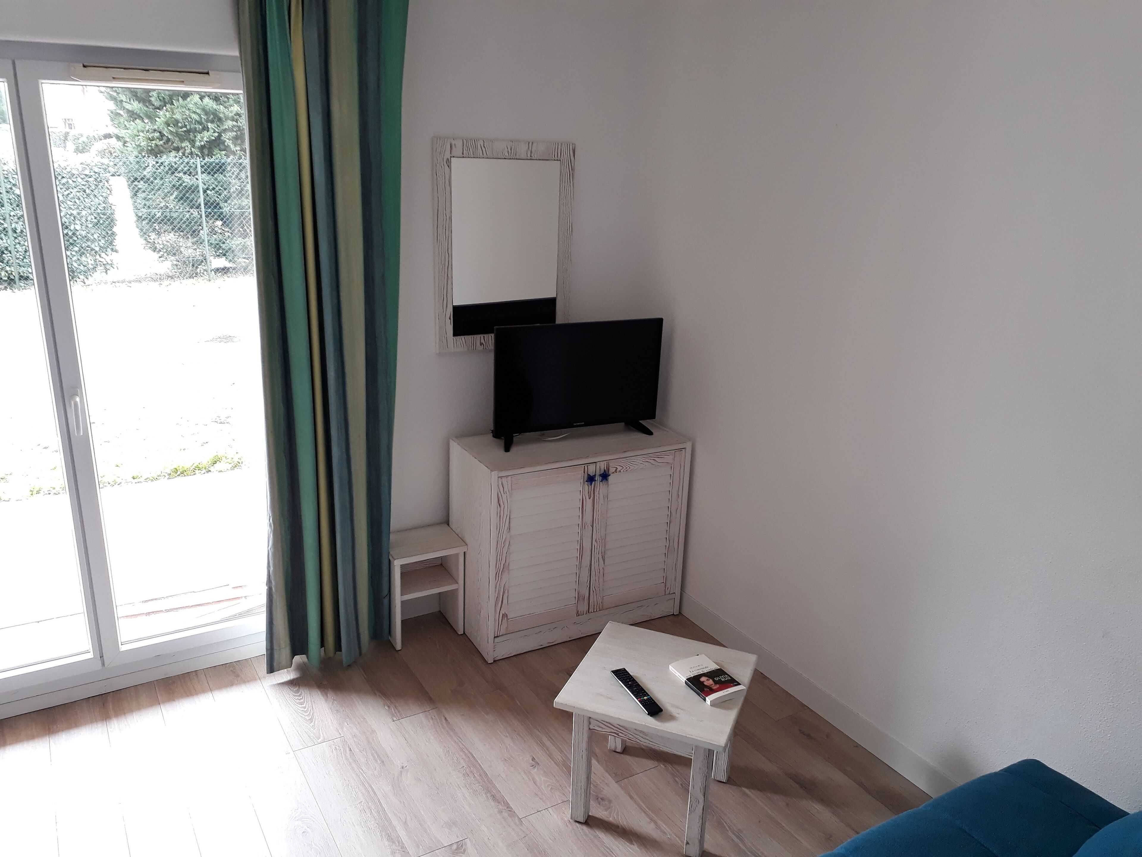 Een kamer met een televisie op een houten kast, een klein wit tafeltje met een afstandsbediening en een boek, en een schuifdeur met gordijnen.