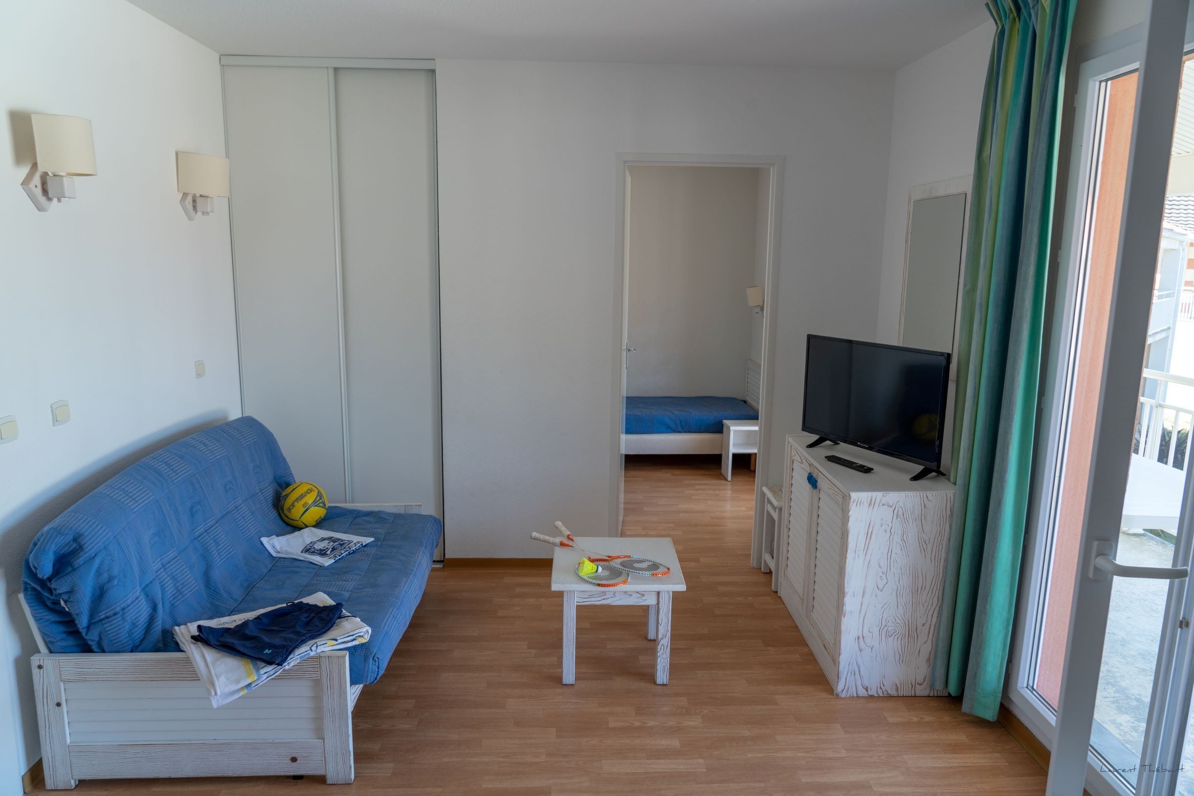 A small, bright room with a blue sofa, a small table with a TV, and a bed in the background.
