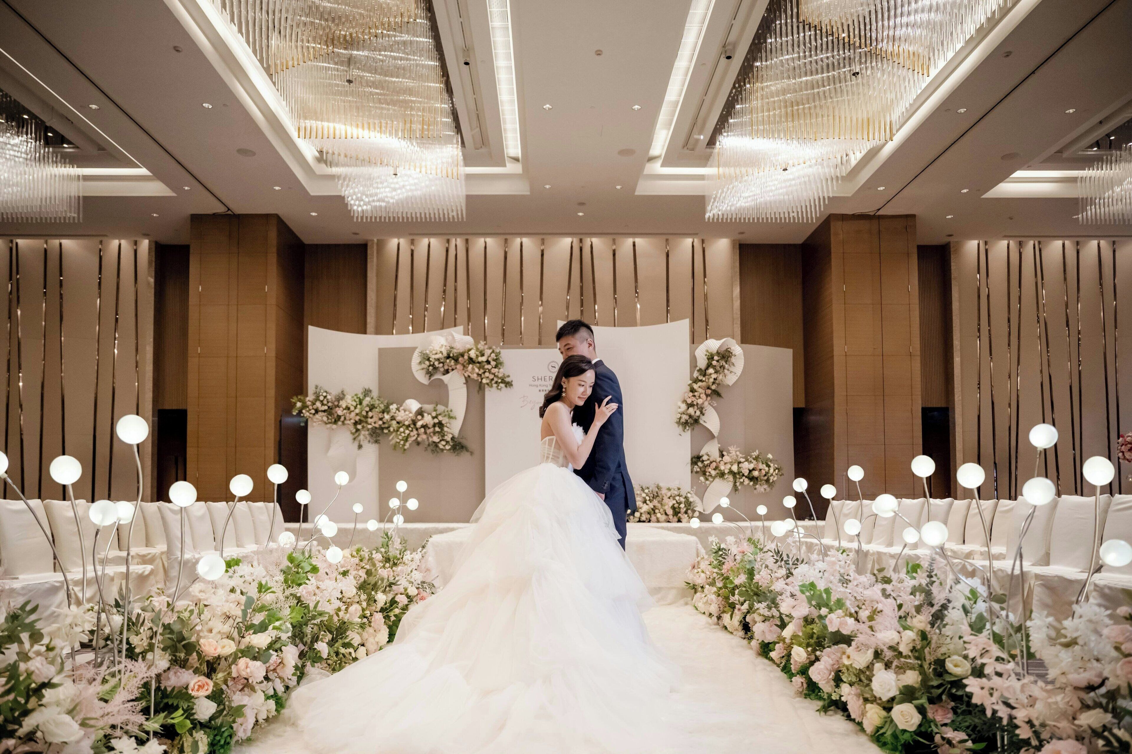 A wedding ceremony with a couple on an aisle decorated with floral arrangements and illuminated by chandeliers.