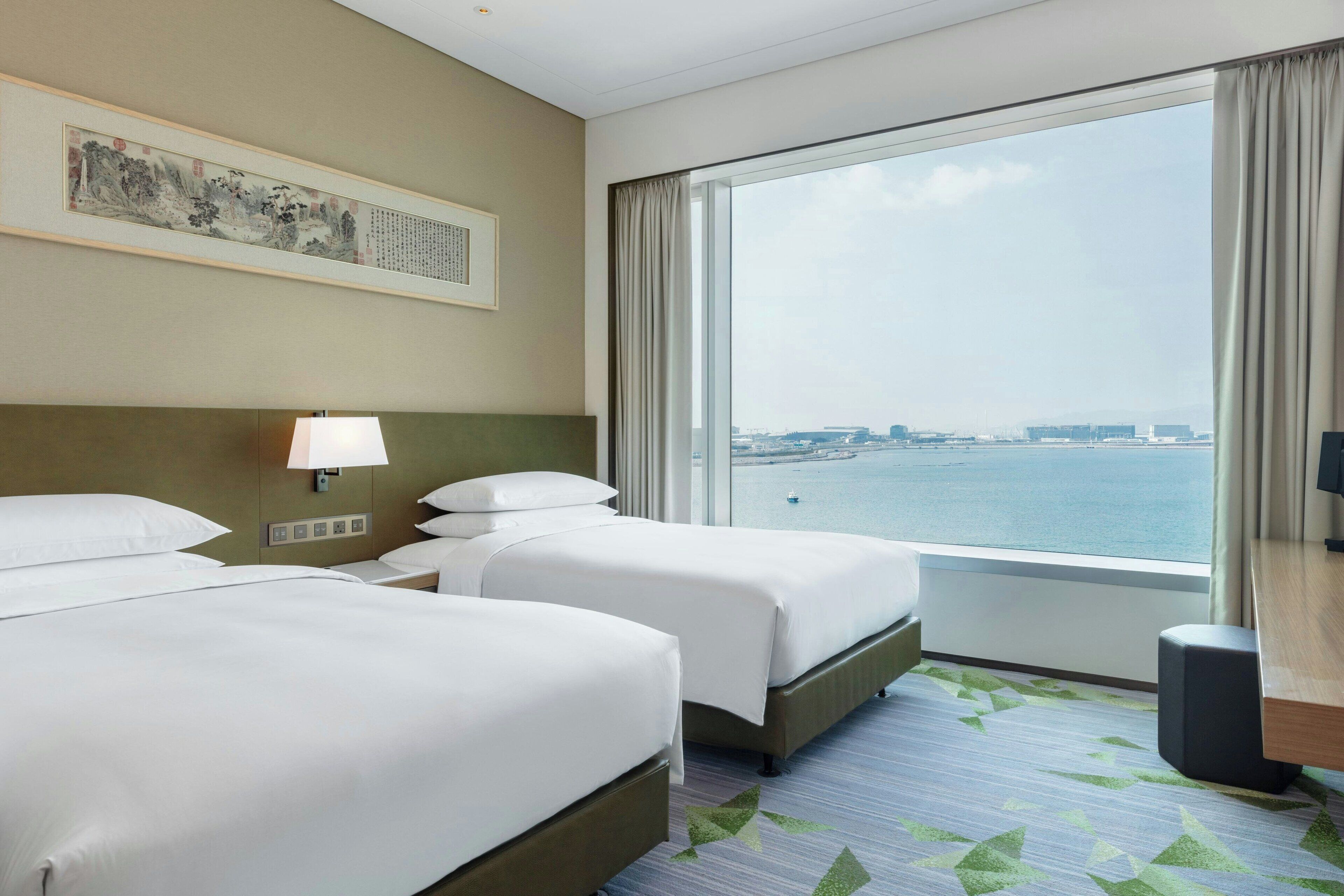 A hotel room with two beds, a large window with a view of the water, a painting on the wall, and a desk with a lamp.