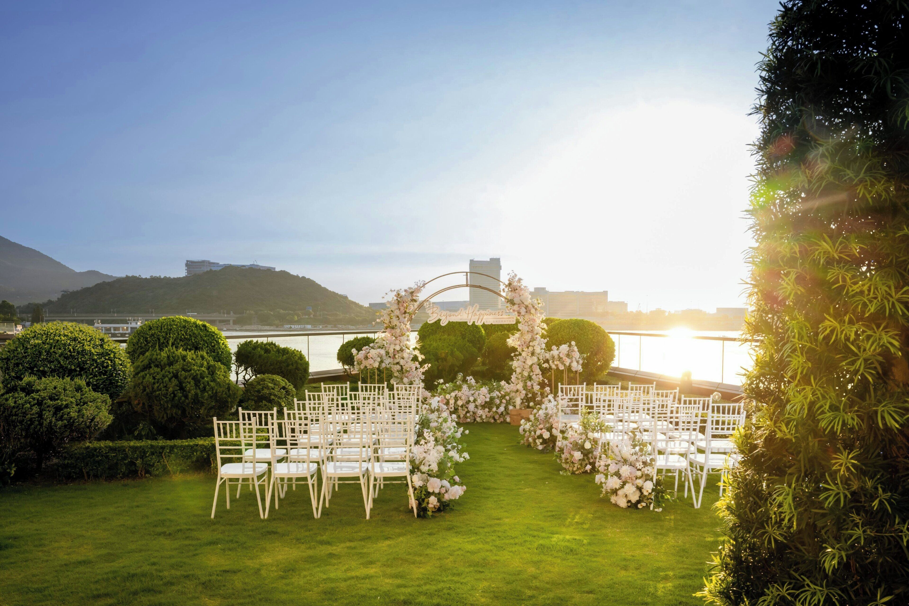 An outdoor wedding venue with white chairs arranged in rows, a floral archway, and a view of a body of water and hills in the background.