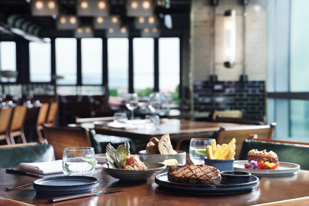 Une table dressée dans un restaurant avec vue, où l’on peut admirer les assiettes remplies de nourriture, les verres et les couverts.