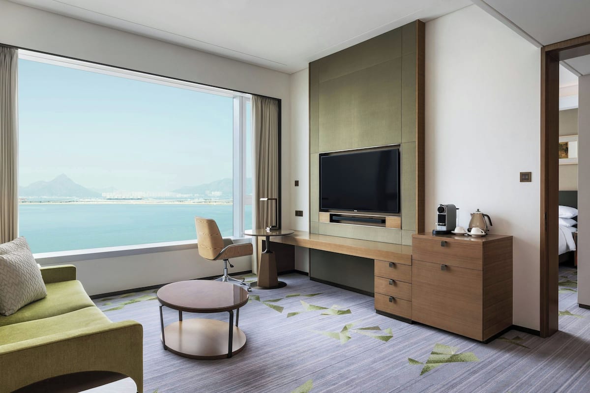 Une chambre d’hôtel dotée d’une grande fenêtre donnant sur la mer et les montagnes, d’un téléviseur à écran plat, d’un canapé, d’un bureau avec une chaise et d’une petite table.