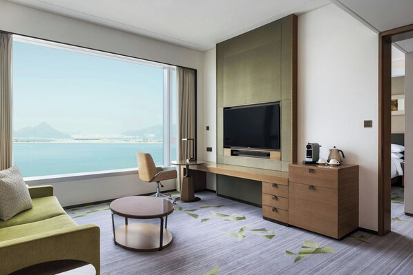 Habitación de hotel con un ventanal que ofrece vistas al mar y a las montañas, un televisor de pantalla plana, un sofá, un escritorio con una silla y una mesita.