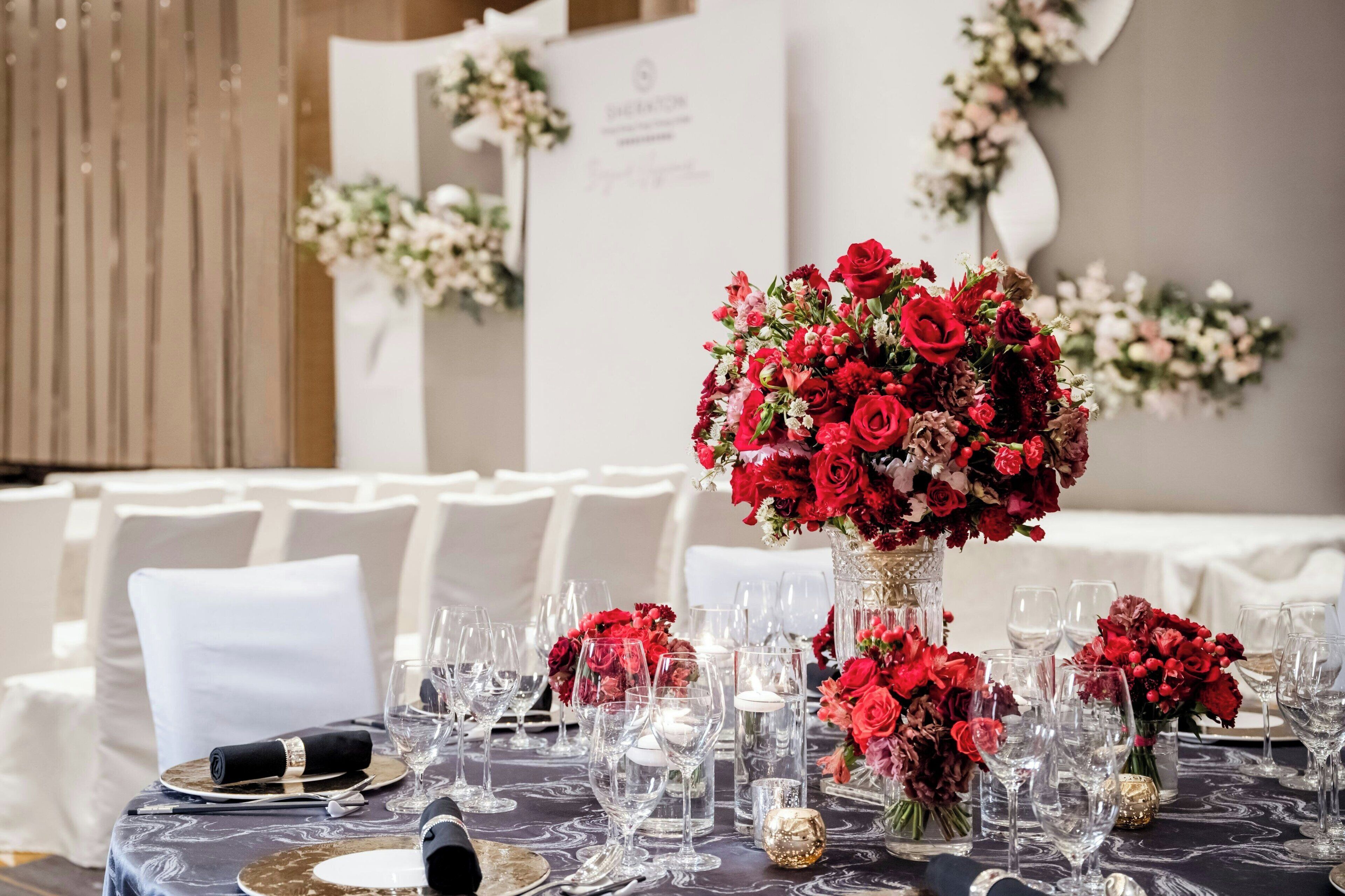 Une table dressée pour un événement formel, agrémentée d’un centre de table composé de fleurs rouges, de chaises blanches et d’un fond blanc.