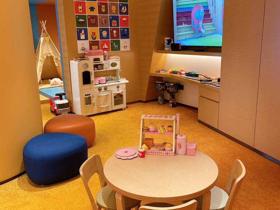 Zona infantil con una mesa redonda, sillas, una cocina de juguete y una pantalla de televisión mostrando un personaje animado.