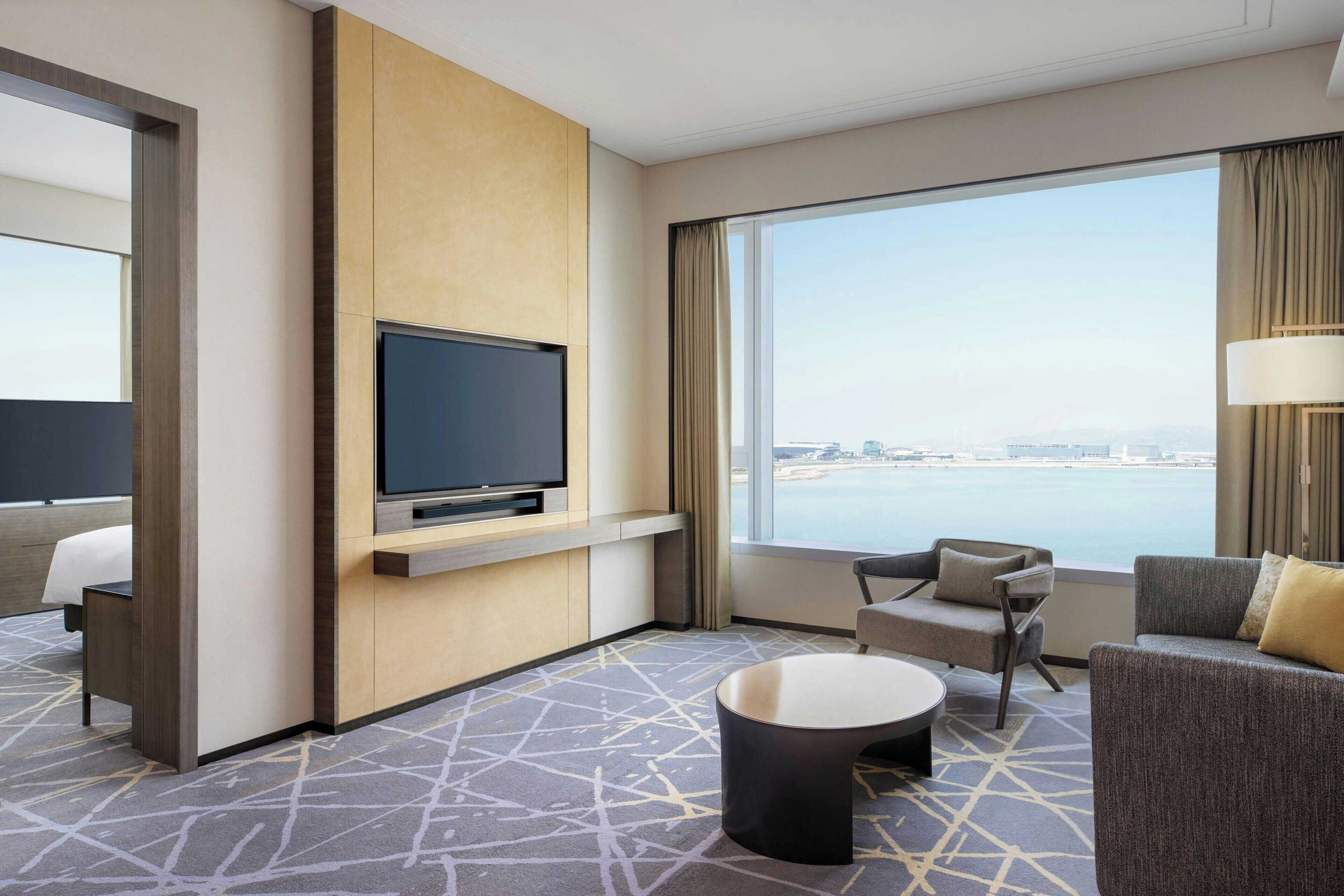 A modern hotel room with a large window, a flat-screen TV mounted on a wooden panel, a sofa, a chair, and a small round table.