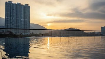 A high-rise building with a sunset reflection on a body of water, a bridge, and a hilly landscape in the background.