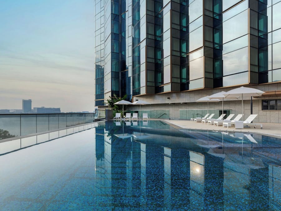 A rooftop infinity pool with lounge chairs and umbrellas, surrounded by a modern building with large windows.
