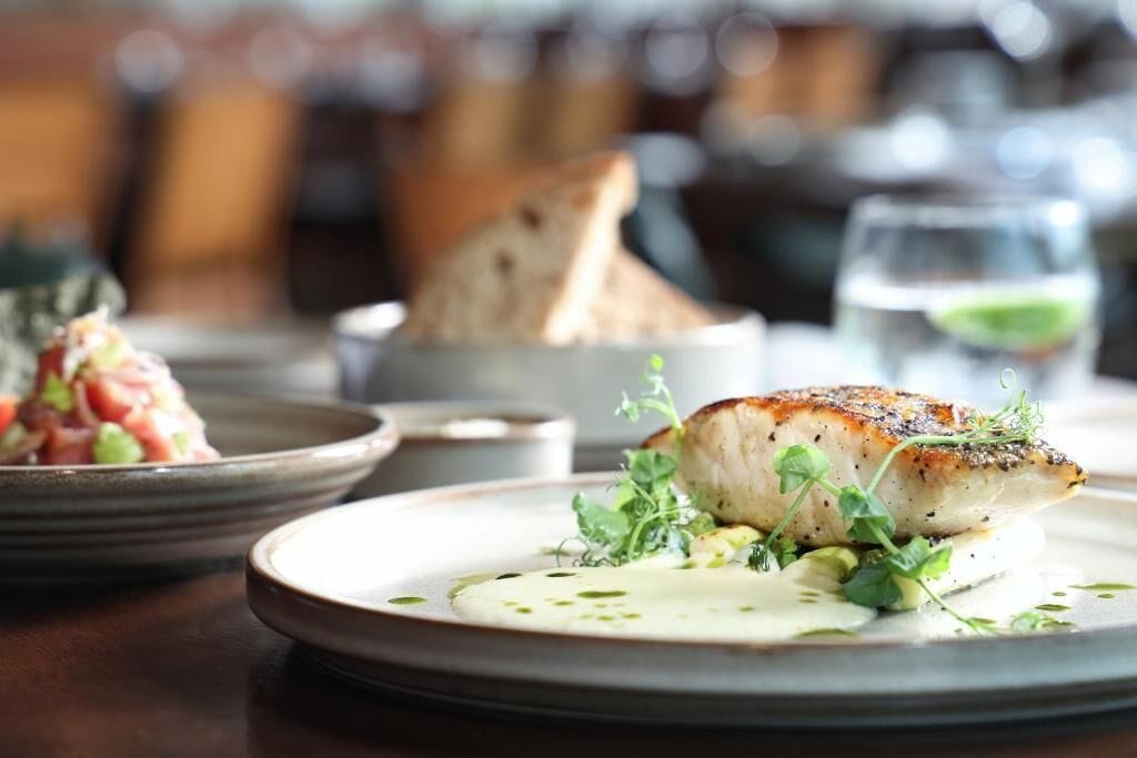 A well-set dining table with a plate of seared fish, a bowl of soup, and a glass of water.