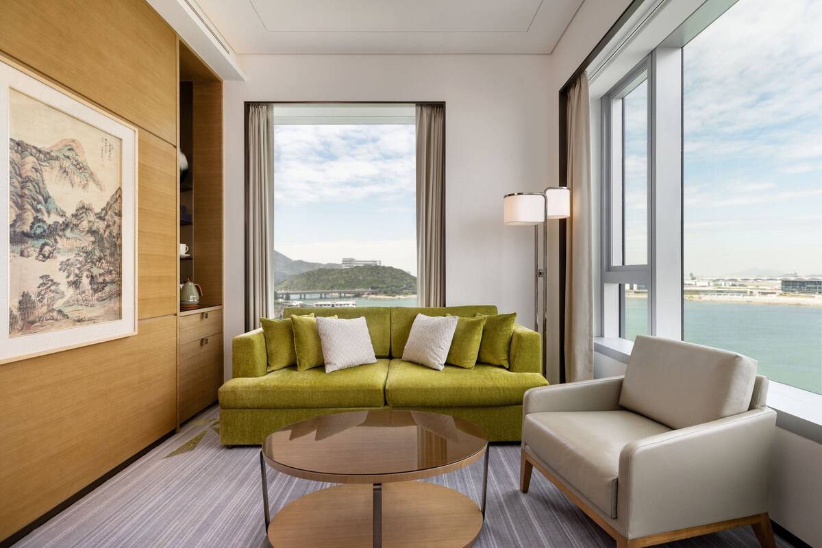 Une chambre d’hôtel moderne avec un canapé vert, un fauteuil beige, une table basse ronde et une grande fenêtre donnant sur la mer et les montagnes.