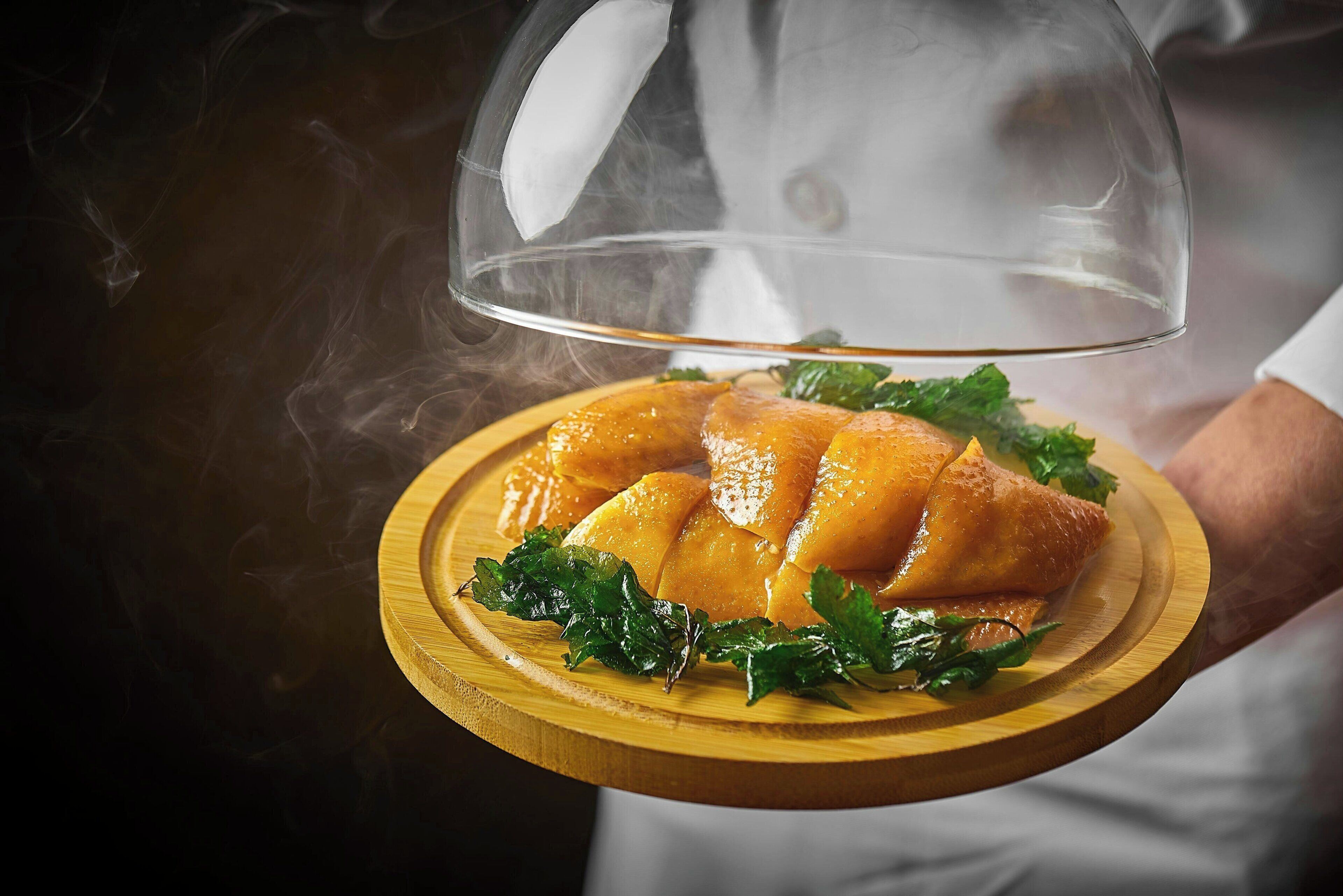 A chef holding a wooden board with a dish covered by a transparent dome, steam rising from the food.