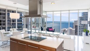 Una cocina moderna con una isla grande, horno empotrado y campana extractora de acero inoxidable. Hay un comedor con una mesa de vidrio y sillas blancas. La habitación tiene ventanas amplias que ofrecen vistas al océano y al horizonte de la ciudad.