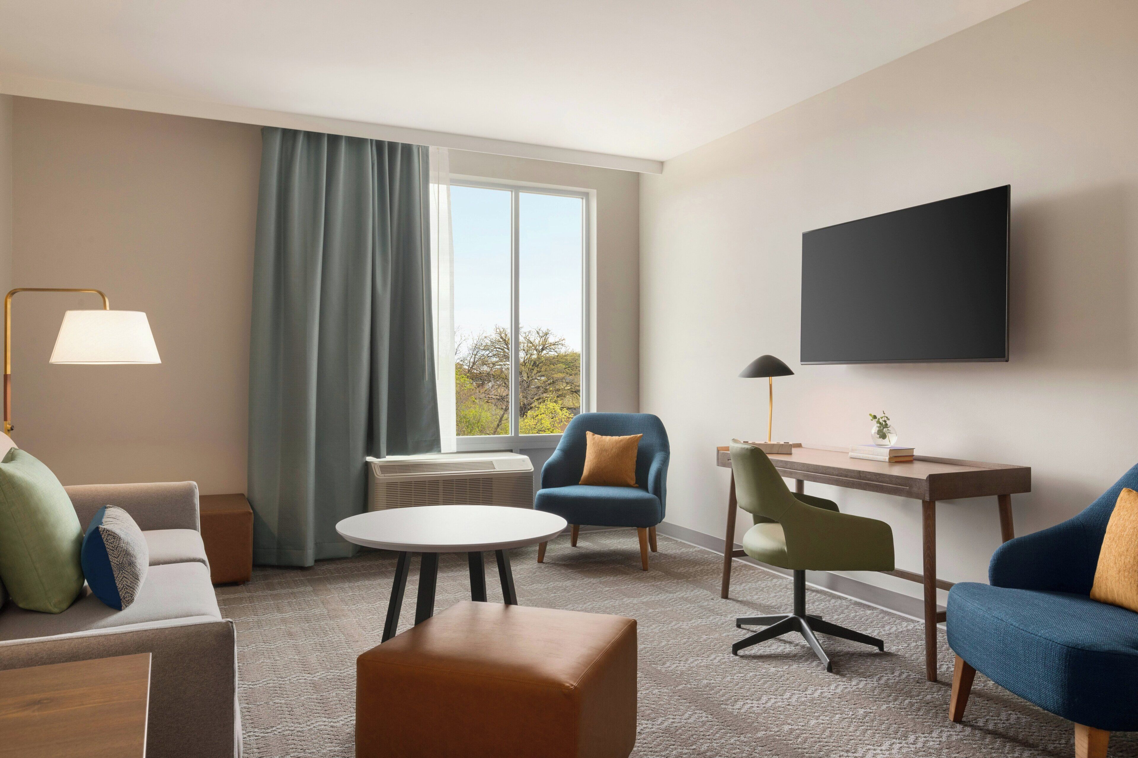 Een hotelkamer met een bank, twee fauteuils, een klein rond tafeltje, een bureau met stoel, een aan de muur bevestigde televisie en een raam met gordijnen.