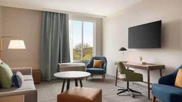 Een hotelkamer met een bank, twee fauteuils, een klein rond tafeltje, een bureau met stoel, een aan de muur bevestigde televisie en een raam met gordijnen.