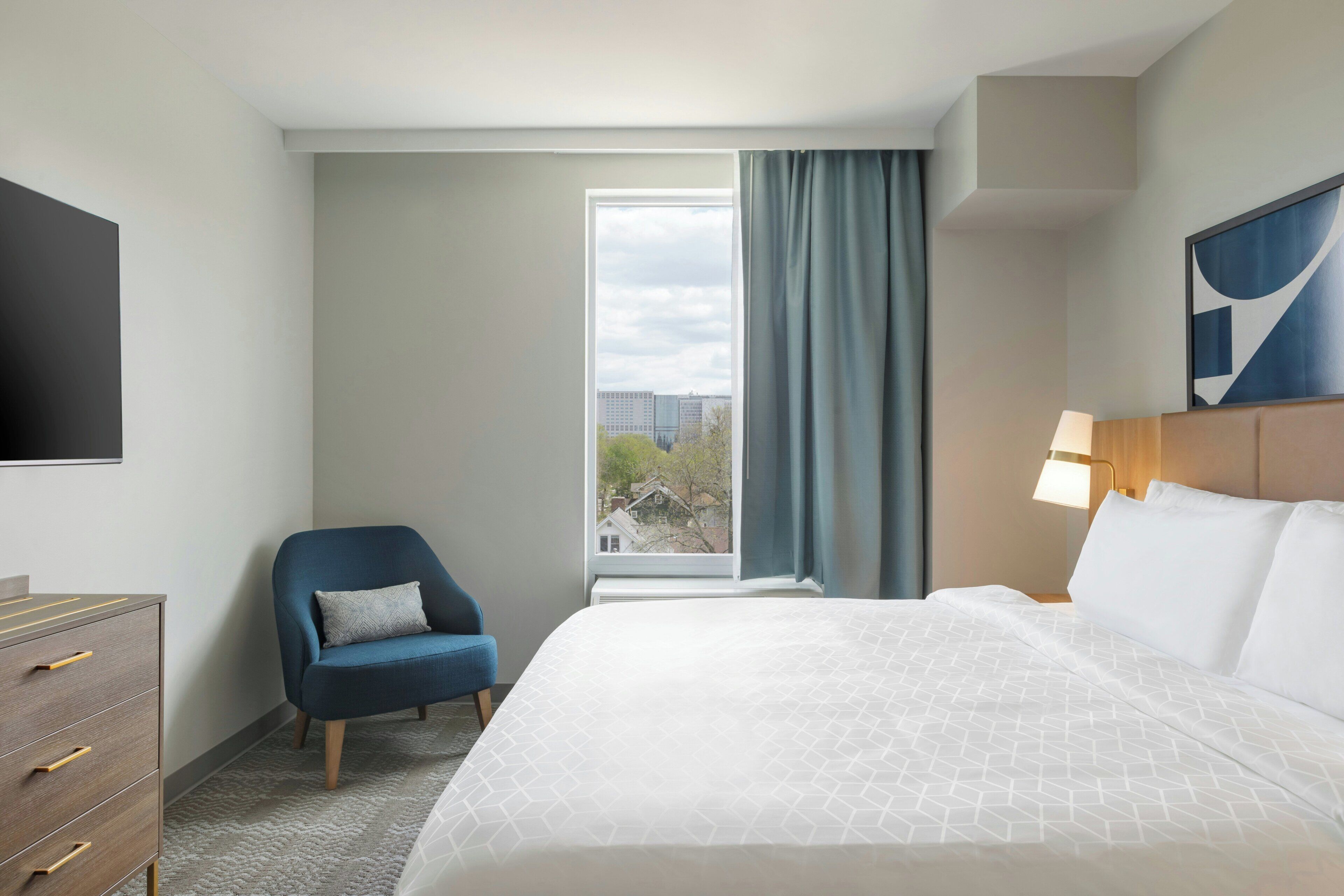 A hotel room with a bed, a chair, a TV, a window with curtains, and a lamp.