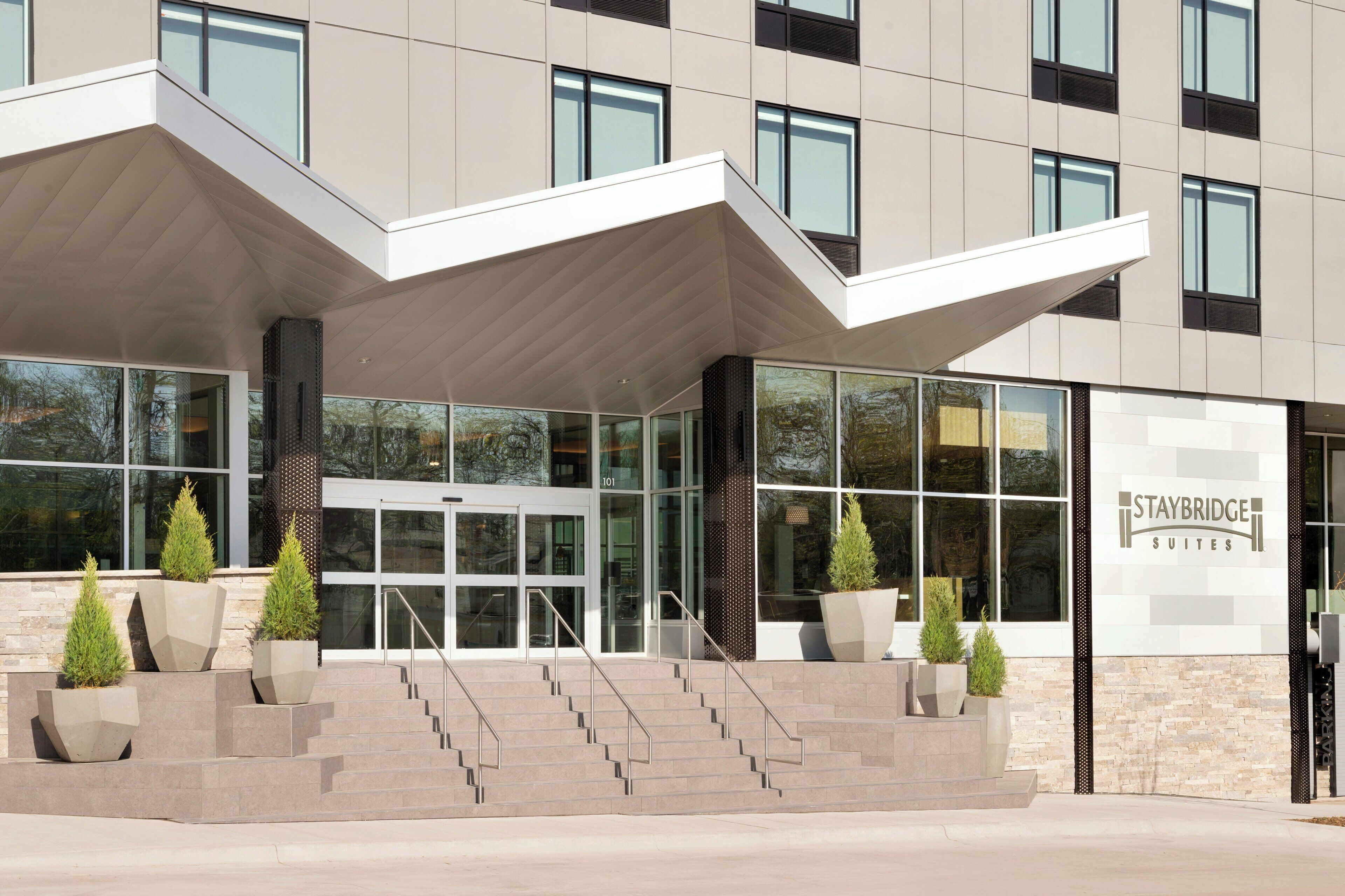 A modern hotel entrance with a glass facade, a staircase leading to the entrance, and a sign for Staybridge Suites.