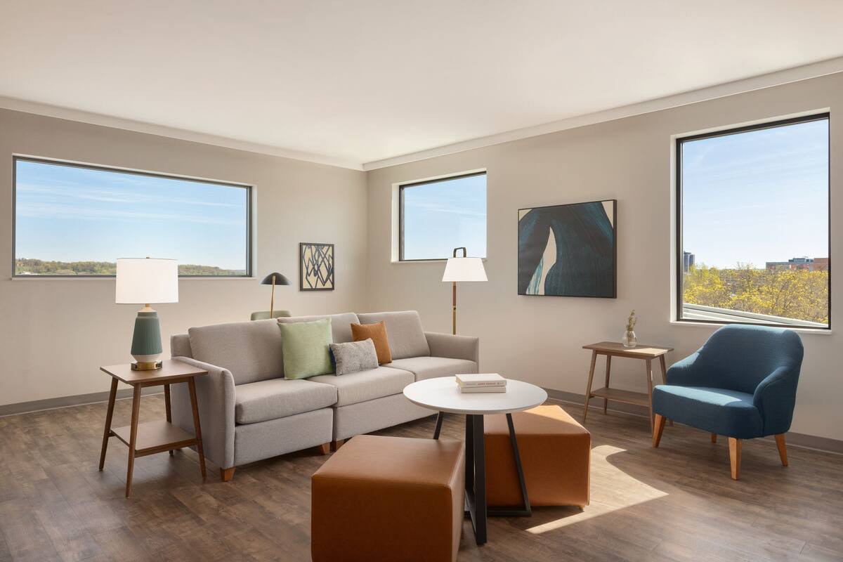 A modern living room with a sofa, armchairs, a coffee table, and a lamp. There are large windows providing views of the outdoors.