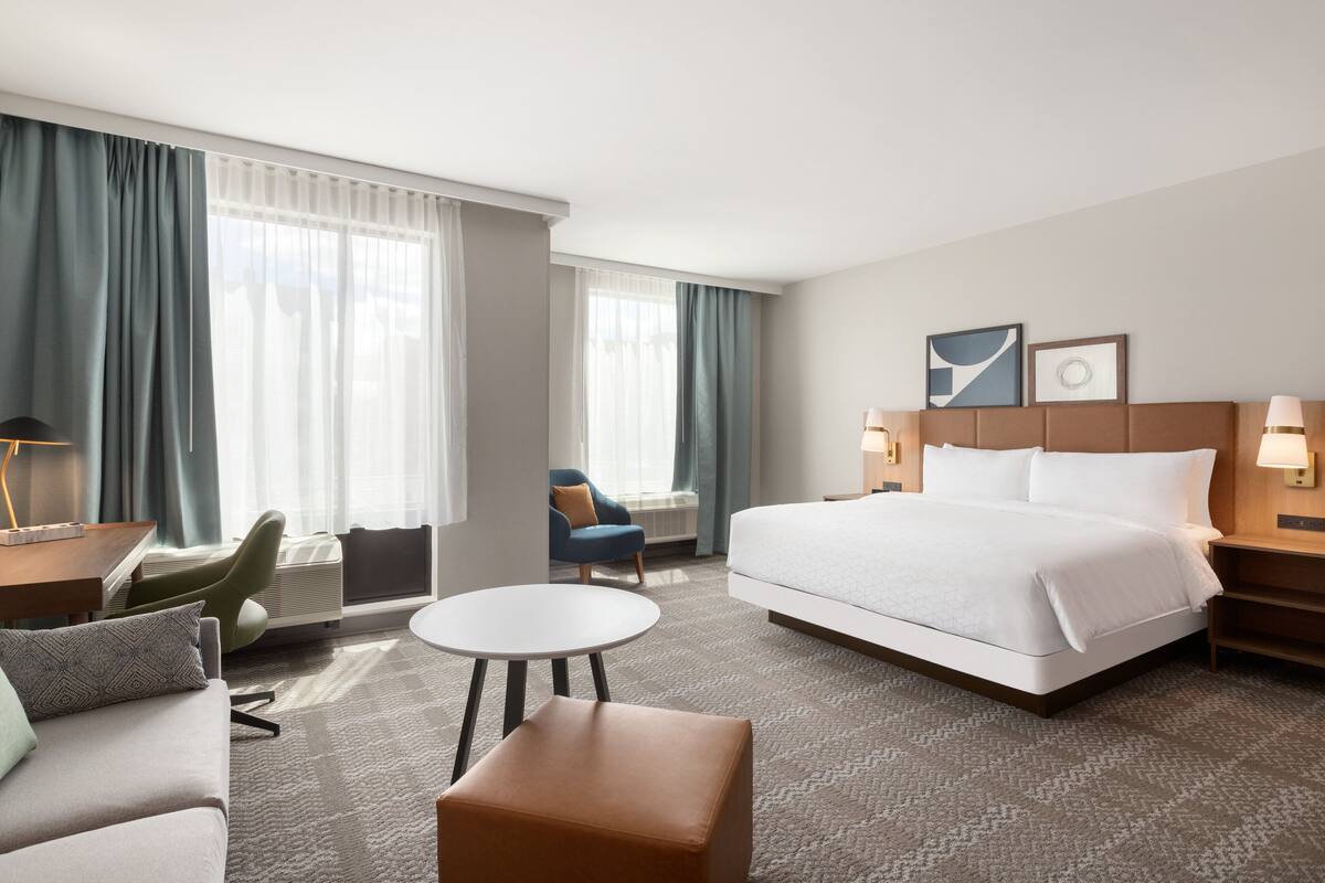 A hotel room with a large bed, a round table, a sofa, and a chair. There are two windows with curtains, a wall-mounted lamp, and a framed picture above the bed.