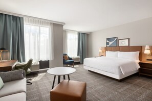 A hotel room with a large bed, a round table, a sofa, and a chair. There are two windows with curtains, a wall-mounted lamp, and a framed picture above the bed.