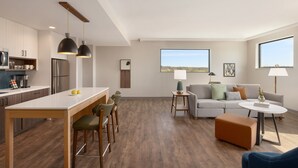 Eine moderne Küche mit einer zentralen Kochinsel, Barhocker und Pendelleuchten. Ein Wohnbereich mit Sofa, Hocker und Blick ins Freie ist sichtbar.