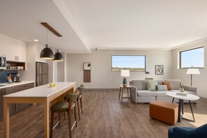 Eine moderne Küche mit einer zentralen Kochinsel, Barhocker und Pendelleuchten. Ein Wohnbereich mit Sofa, Hocker und Blick ins Freie ist sichtbar.