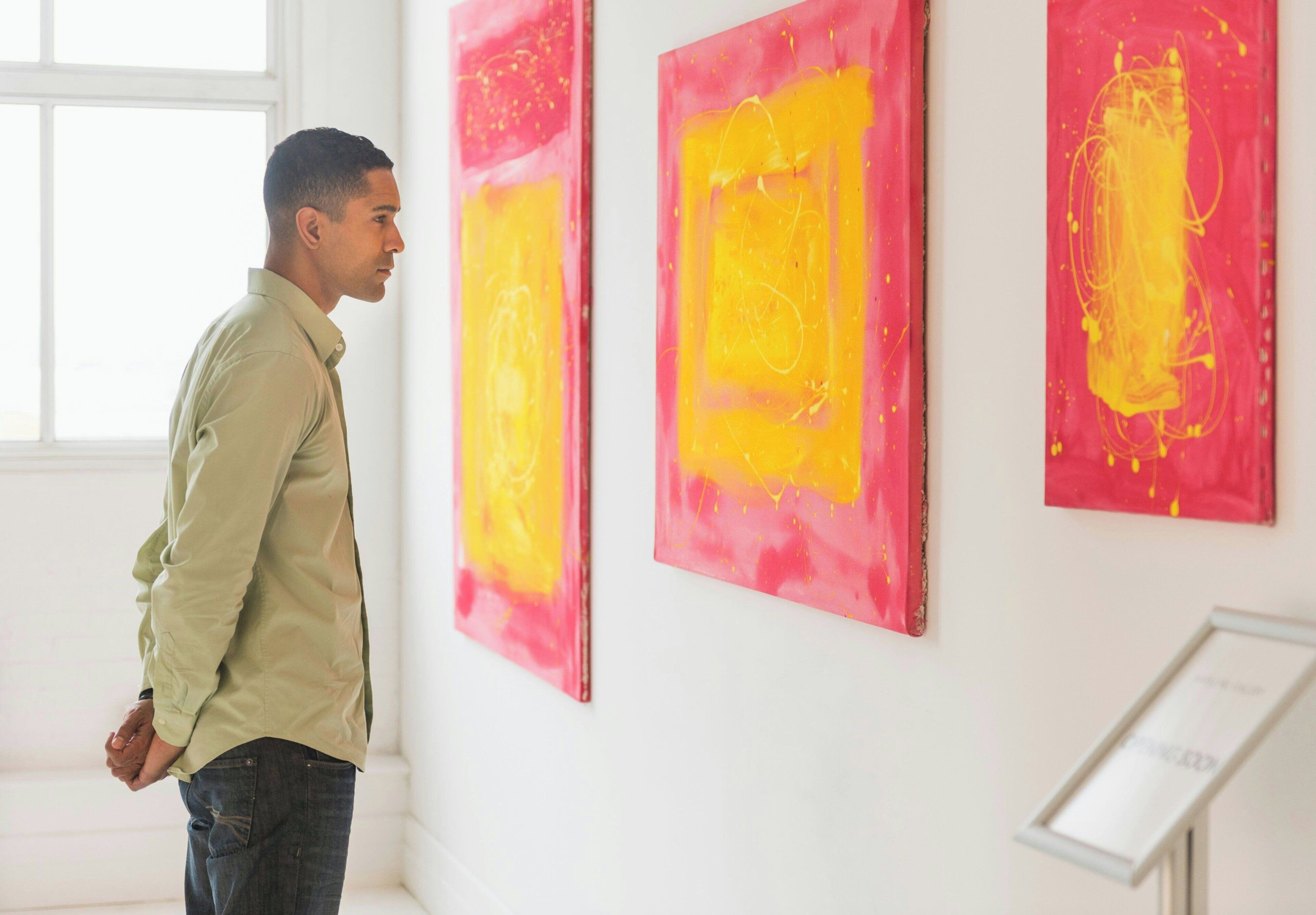 A person standing in a room with white walls and a window, observing paintings with red and yellow colors.