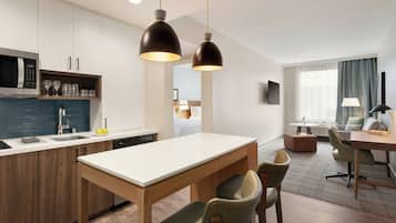 A modern hotel room with a kitchen area, a dining table with chairs, a sofa, and a TV mounted on the wall.