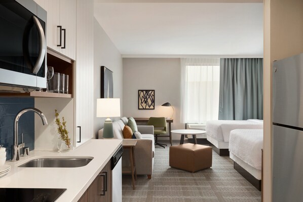 A hotel room with a kitchenette, a bed, a sofa, a small table, and a window with curtains.