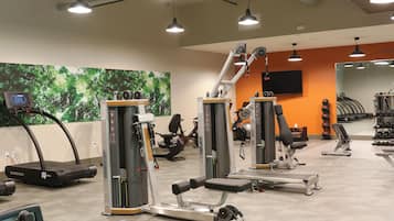 Une salle de sport équipée de divers appareils d’exercice, d’un tapis de course et d’un écran fixé au mur.