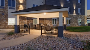 A covered outdoor patio with seating and a barbecue area, adjacent to a multi-story building with stone and brick facade. - Candlewood Suites San Antonio Schertz by IHG (Schertz)
