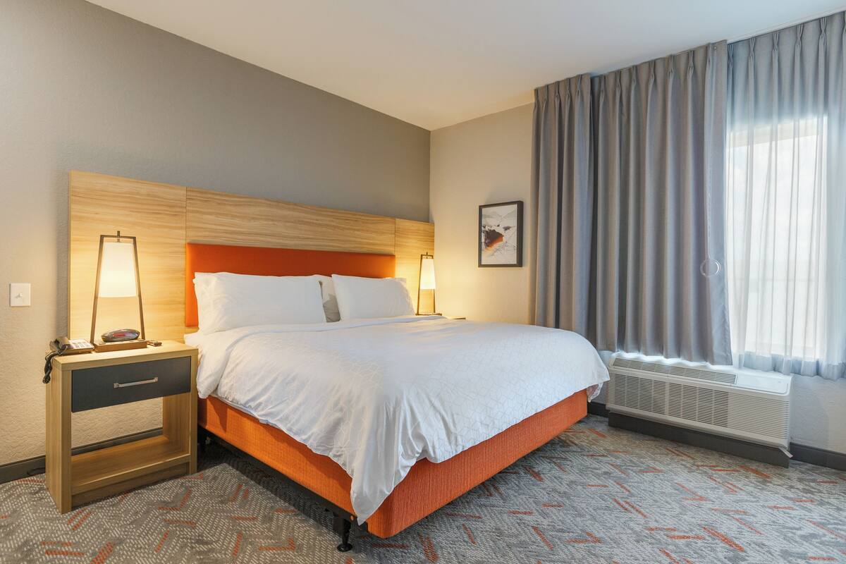A hotel room with a bed, nightstand, lamp, and a window with curtains.