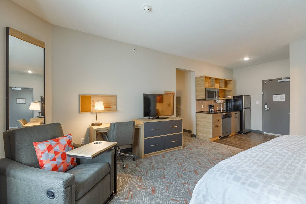 A hotel room with a bed, a desk, a chair, a sofa, a TV, a microwave, a refrigerator, and a small kitchenette.
