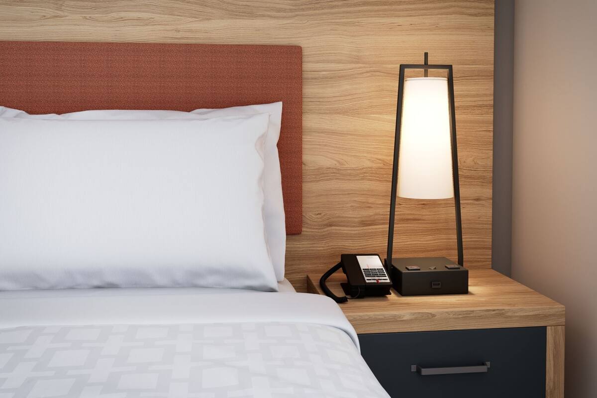 A neatly made bed with white linens, a wooden nightstand with a lamp, and a drawer.