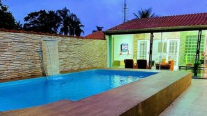 A swimming pool with a wooden deck, a small waterfall feature, and a covered seating area with chairs and tables.