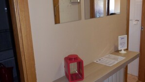 A hallway with a wooden floor, a white wall with a mirror and a red object on a shelf, and a door.