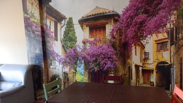 A dining area with a table and chairs, a grey sofa, and a mural of a street scene with buildings and purple flowers.
