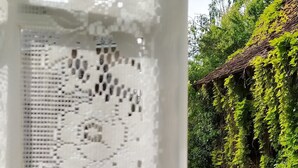 Uma janela de renda branca com vista para um jardim verdejante e um edifício rústico.