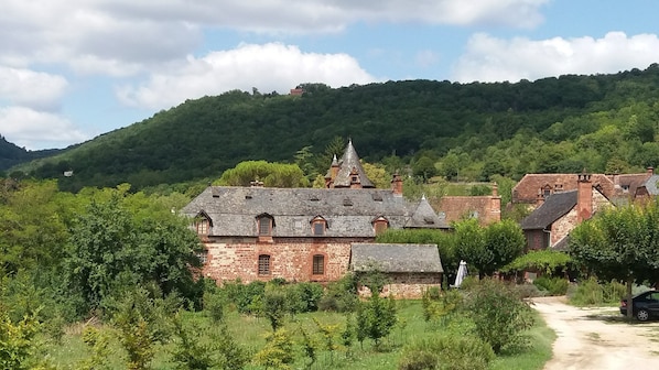 A stone building with a steep roof, surrounded by lush greenery and a forested hill in the background.