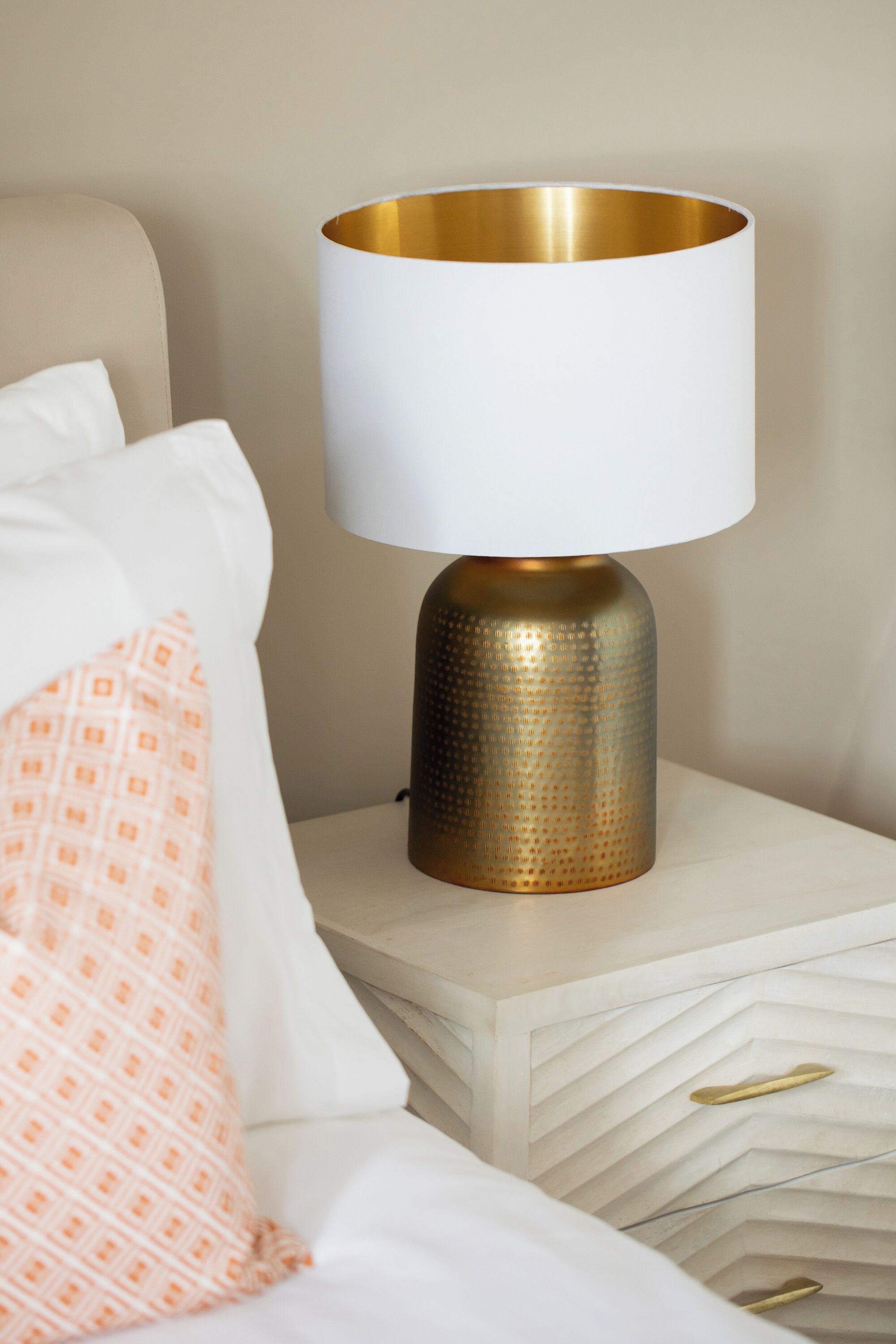 A bedside table with a lamp, a bed with white and peach pillows, and a white nightstand with drawers.