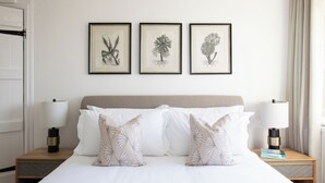 A neatly made bed with white linens and decorative pillows, flanked by two bedside tables with lamps and books. Above the bed, three framed botanical illustrations are hung on the wall. - Beautiful Iconic Listed Building with Views Overlooking the Arundel Cathedral (Arundel)