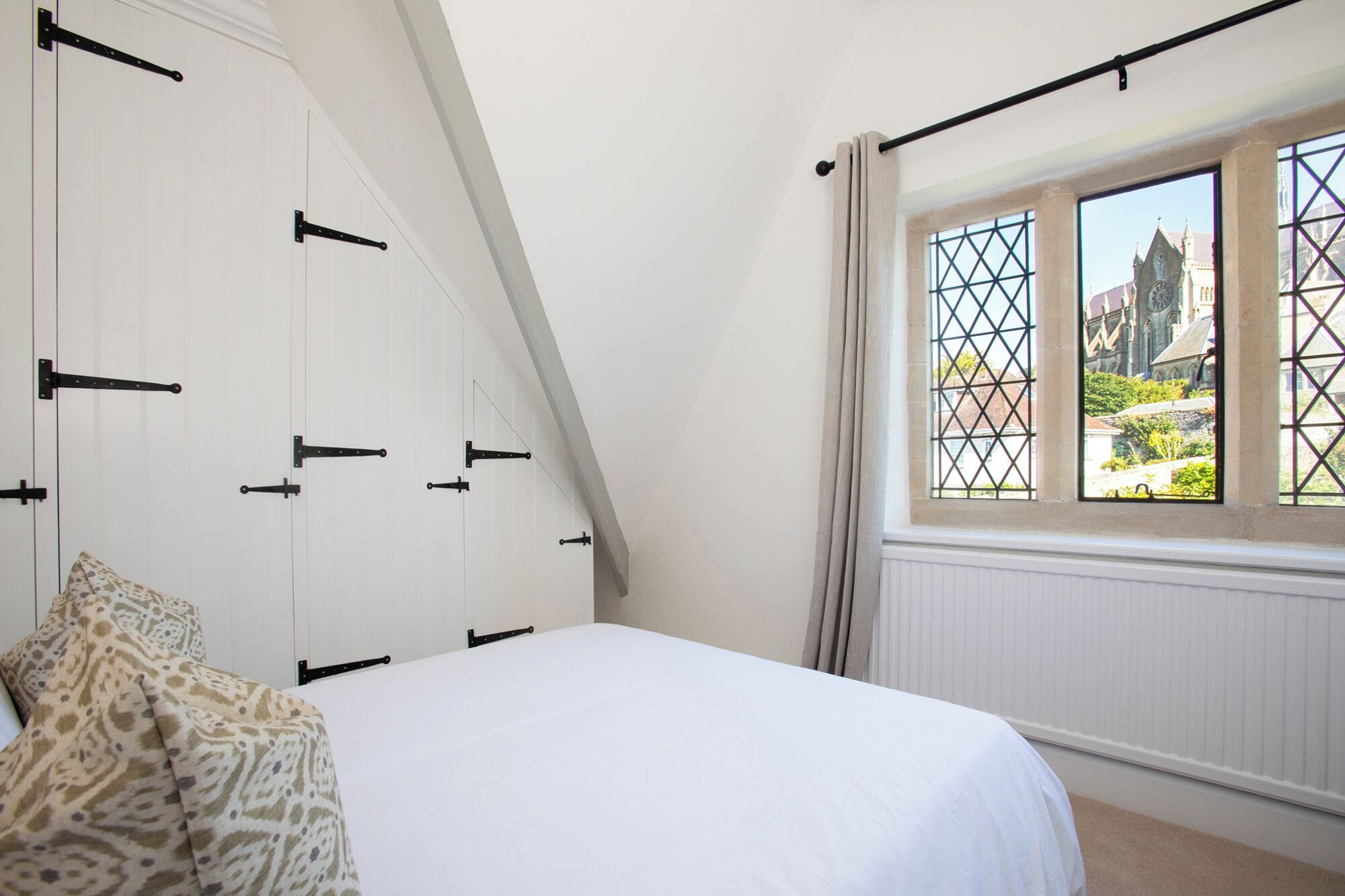 A bedroom with a bed, a window with a view of buildings, and a white wall with a black metal structure.